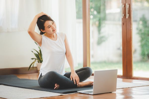 Frau sitzt auf einer Matte vor einem Laptop und macht eine Dehnübung für den Nacken.