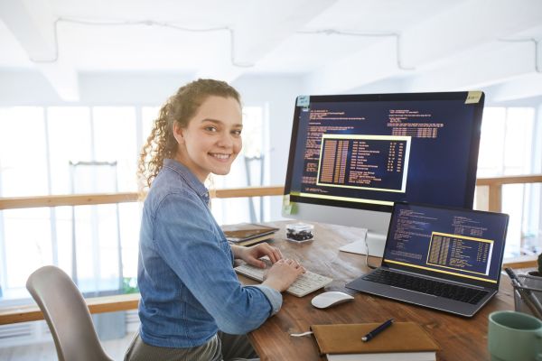 Beleuchtung am Arbeitsplatz. Frau sitzt am Schreibtisch, arbeitet am Computer mit Monitor und Laptop und schaut lächelnd zur Kamera.