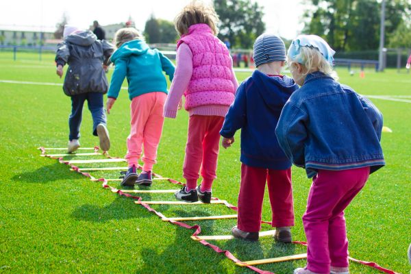 Kinder balancieren draußen über eine Koordinationsleiter auf dem Sportplatz und bewegen sich gemeinsam im Freien.