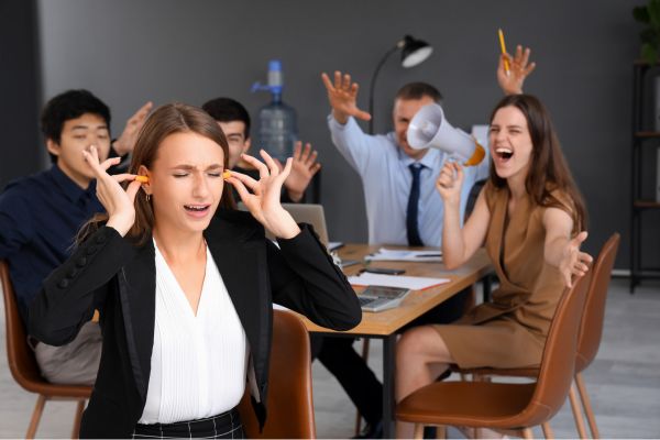 Frau hält sich im Büro die Ohren zu, während mehrere Personen im Hintergrund laut gestikulieren und sprechen.